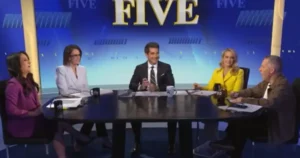 A group of five people are seated around a table in a news studio, engaged in a discussion.