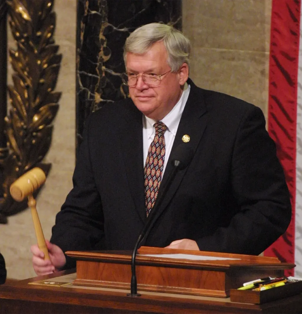 A man in a suit and tie is standing at a podium, holding a gavel, and appears to be presiding over a formal event or meeting.