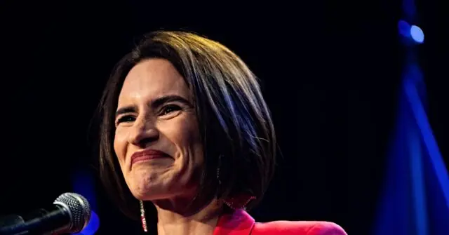 The image shows a person speaking at a podium with a microphone, wearing a red outfit, and standing in front of a dark background with a blue light effect.