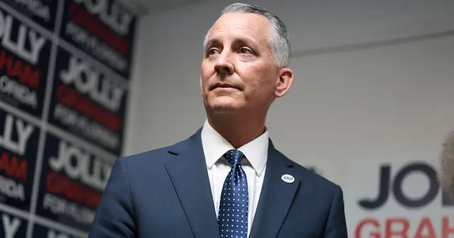 The image shows a man in a suit and tie standing in front of campaign signs, suggesting a political event or campaign setting.