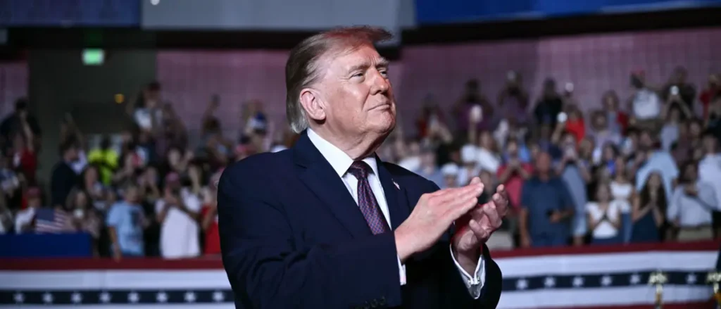 The image shows a man in a suit clapping in front of a crowd at what appears to be a political rally.