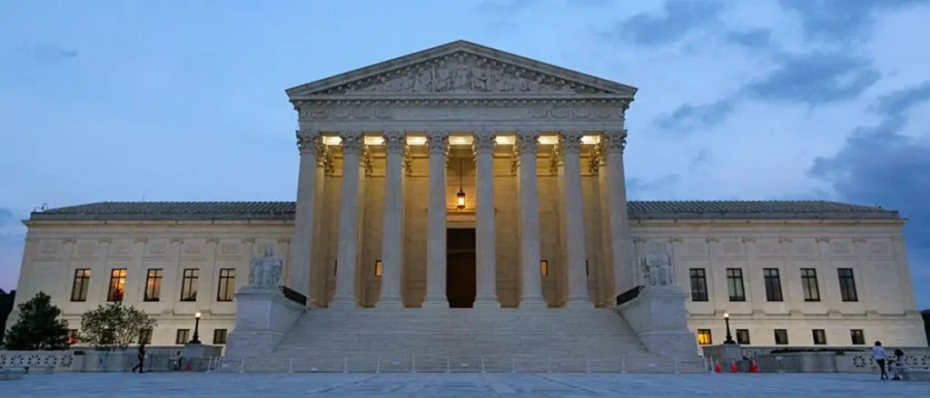 The image shows the United States Supreme Court building at dusk, with its iconic columns and steps illuminated against a twilight sky.