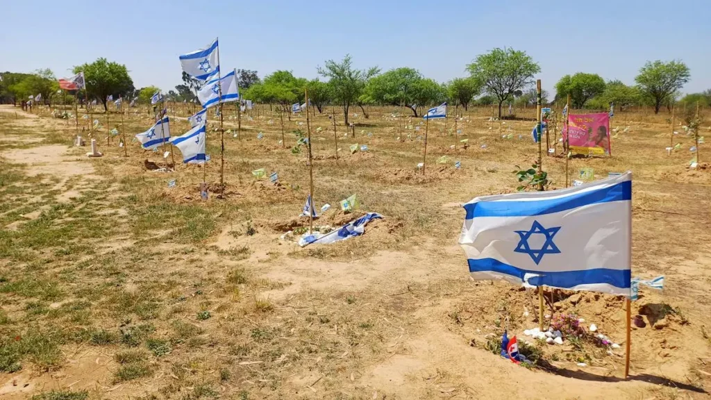 The image shows a memorial site with numerous Israeli flags planted in the ground, likely commemorating fallen soldiers or victims.