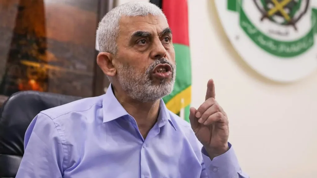 The image shows a man in a light blue shirt gesturing with his right hand, with a flag and emblem in the background.