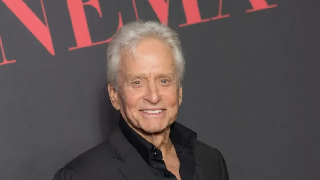 The image shows a person smiling at the camera, dressed in a black suit, with a backdrop featuring red text.