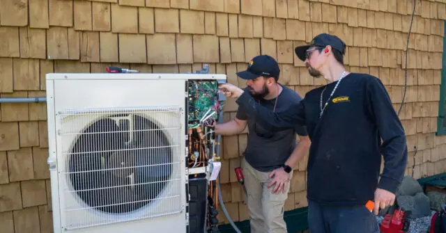Two individuals are examining an outdoor HVAC unit, pointing at its internal components.