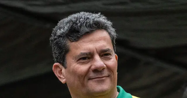 The image shows a person with short, curly gray hair, wearing a green and yellow jacket, standing in front of a dark, textured background.