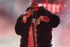The image shows a performer on stage, wearing a fur coat and a red shirt, holding a microphone and singing.