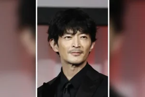 The image shows a man in a black suit and tie, smiling and looking directly at the camera.