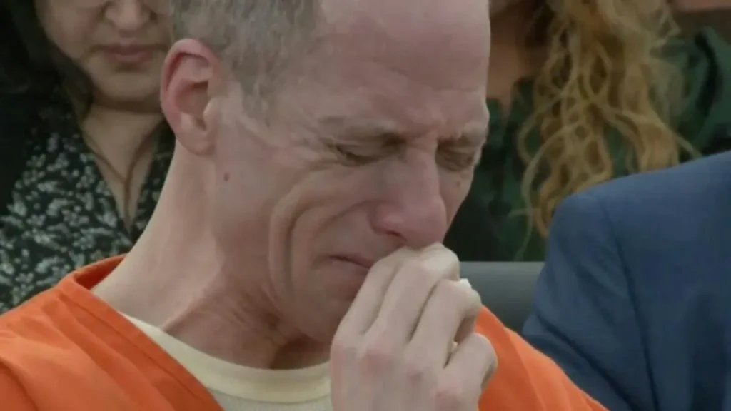 The image shows a man in an orange prison uniform sitting in a courtroom, appearing contemplative with his hand on his chin.