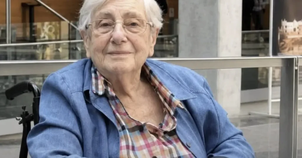 The image shows an elderly person sitting in a wheelchair, wearing a plaid shirt and a denim jacket, with a background that includes a glass railing and some architectural elements.