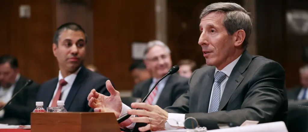 A man in a suit is speaking at a formal meeting or hearing, gesturing with his hands as he addresses the audience.