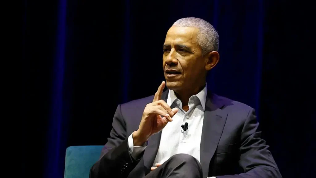 The image shows a man in a suit sitting on a stage, gesturing with his hand as he speaks.