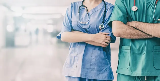 Two healthcare professionals, one in blue scrubs and the other in green scrubs, stand with their arms crossed in a medical setting.