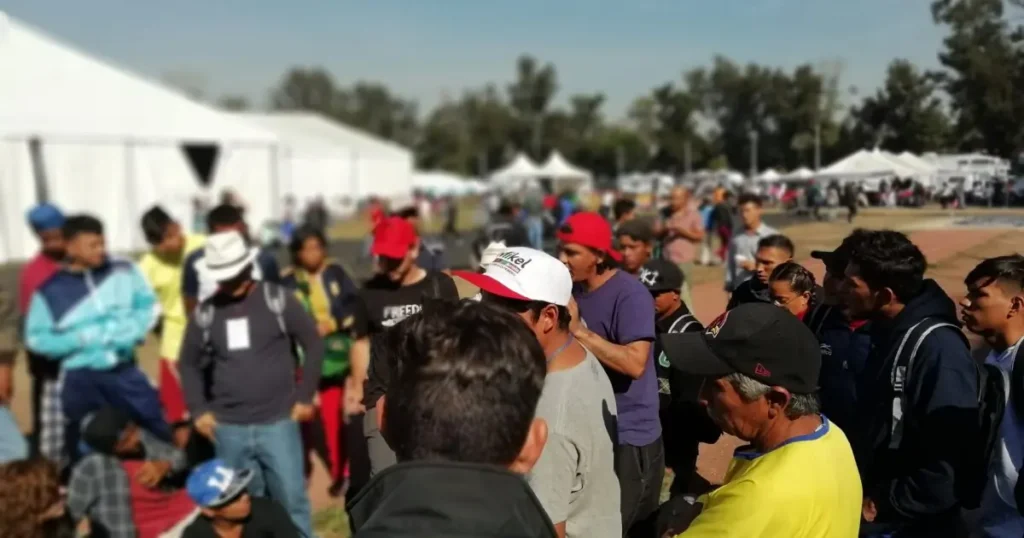 The photo depicts a large group of people gathered outdoors, possibly at a community event or festival, with tents and trees in the background.