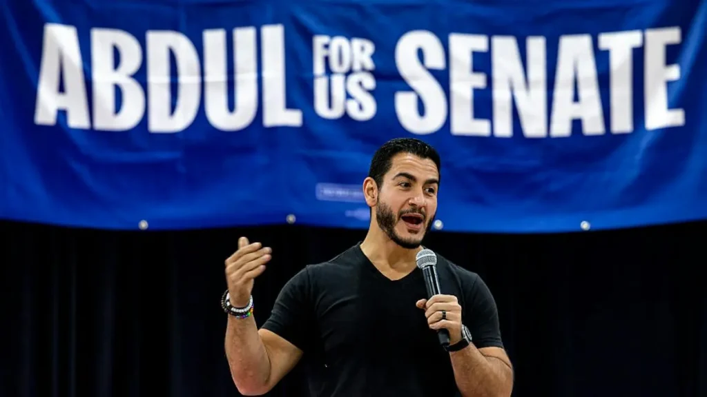 A man is speaking into a microphone in front of a banner that reads "ABDUL FOR US SENATE."