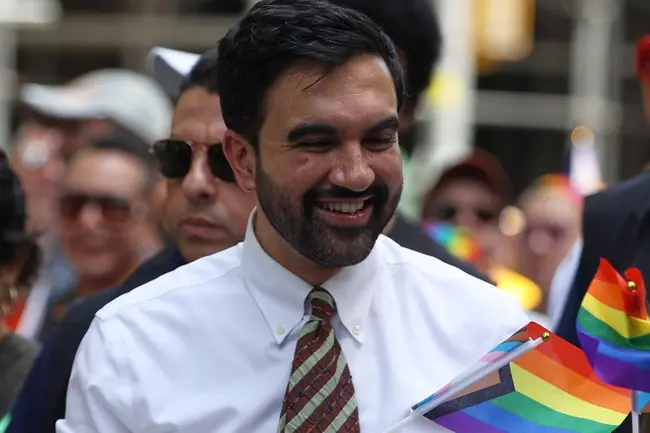 A man smiles while holding a rainbow flag, participating in a pride parade.