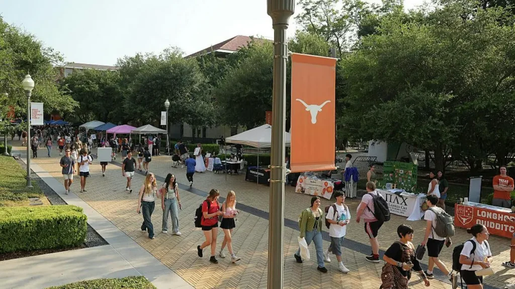 The photo captures a lively scene on a university campus with students walking and interacting near various tents and banners, including one featuring the Longhorn logo.