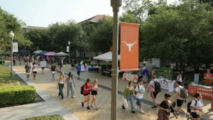 The photo captures a lively scene on a university campus with students walking and interacting near various tents and banners, including one featuring the Longhorn logo.