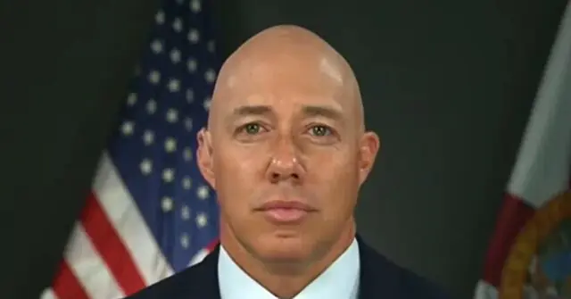 The image shows a person in formal attire with an American flag in the background, suggesting a formal or official setting.