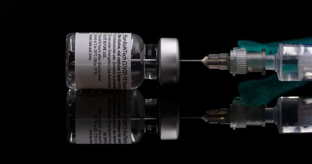 The image shows a vial of Pfizer-BioNTech COVID-19 vaccine being prepared for injection.