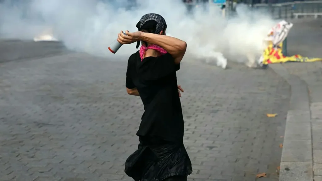 A protester is seen from behind, holding a canister, as smoke billows in the background during a demonstration.