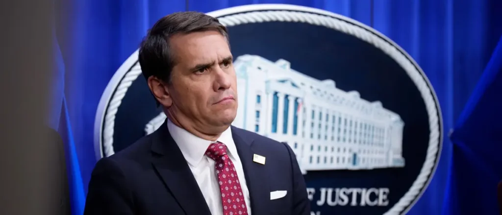 The image shows a man in a suit and tie standing in front of a backdrop featuring the U.S. Department of Justice seal.