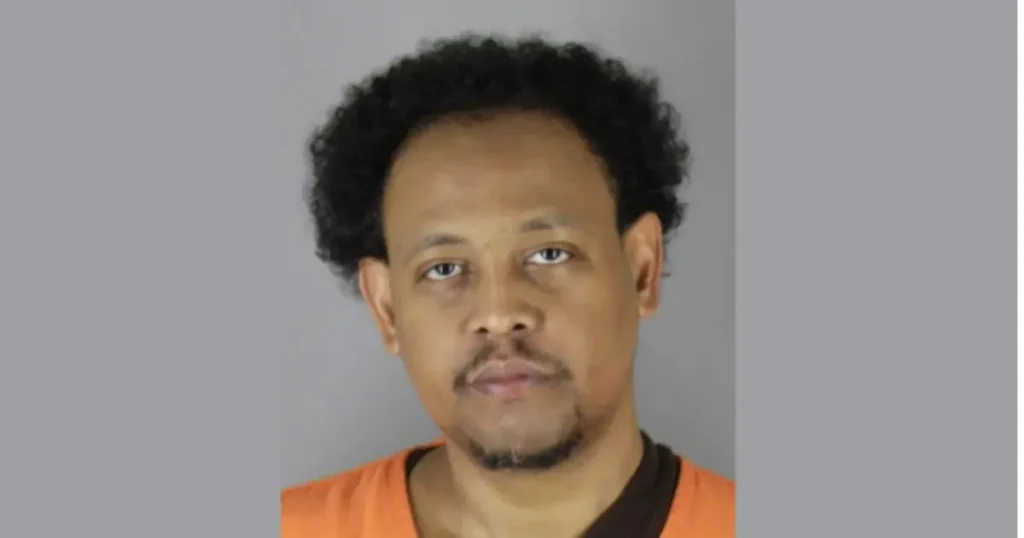 The image is a mugshot of a man with curly hair, wearing an orange shirt.