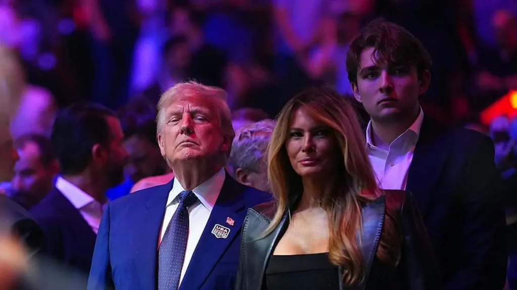 The photo shows a group of people, including a man in a suit and tie, a woman in a black dress, and another man in a suit, standing together in a crowd with a purple-lit background.