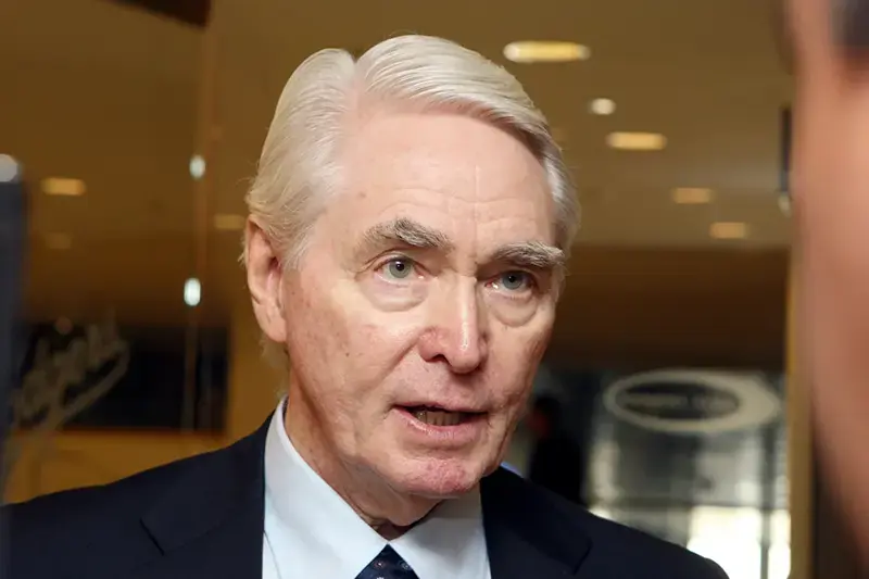 The image shows a man in a suit and tie, speaking in what appears to be a professional or formal setting.