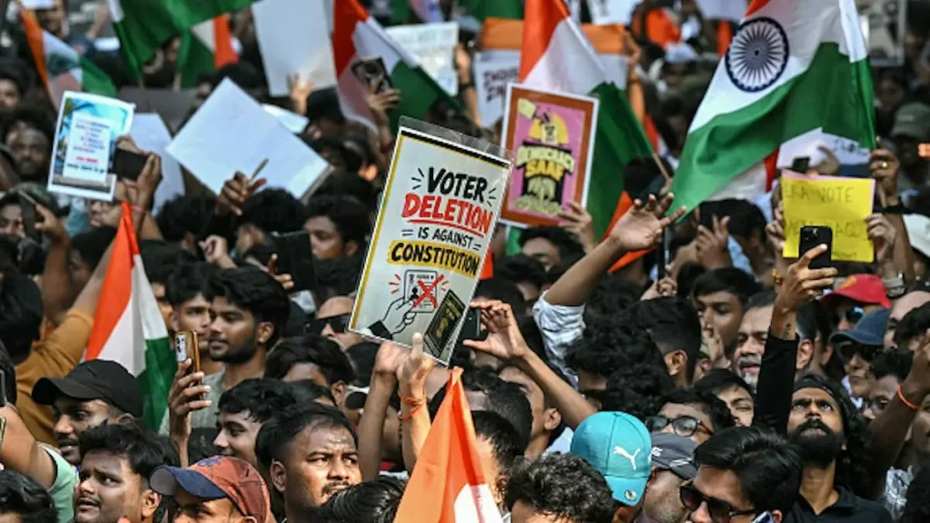 A large crowd of people holds up signs and flags, with one sign reading "Voter Deletion is Against Constitution," indicating a protest or demonstration.