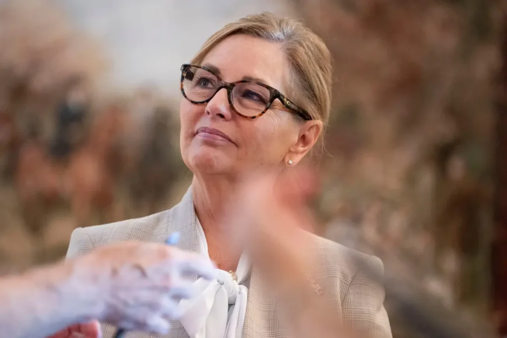 A woman in a professional setting, wearing glasses and a suit, appears to be engaged in a discussion or presentation.