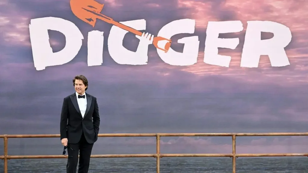 A man in a tuxedo stands in front of a backdrop featuring the word "DIGGER" with a graphic of a shovel.
