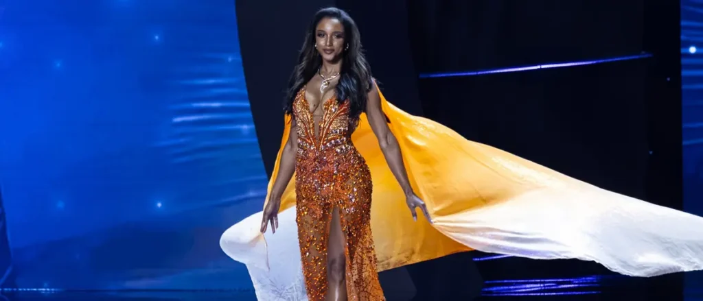 A contestant in a sparkling orange gown with a flowing cape walks the runway during a pageant.