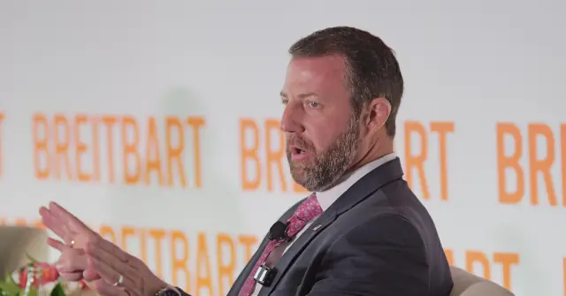 A man in a suit and tie is speaking at a podium with the word "Breitbart" repeated in the background.