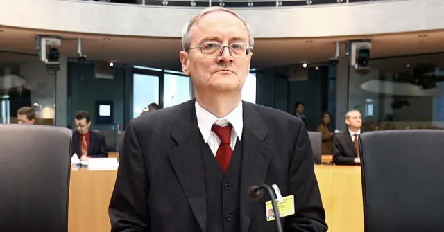 The image shows a man in formal attire standing in what appears to be a conference or parliamentary setting, with microphones and other individuals seated in the background.
