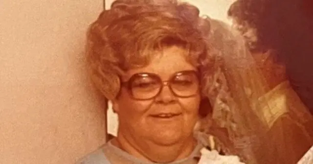 The image shows an elderly woman with curly hair and glasses, smiling and wearing a light-colored top.