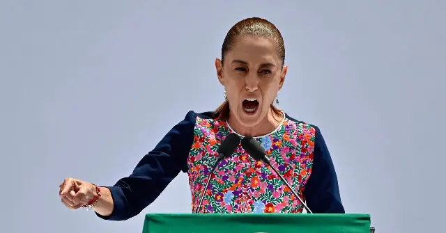 A woman stands at a podium, passionately speaking with her fist raised, against a clear sky.