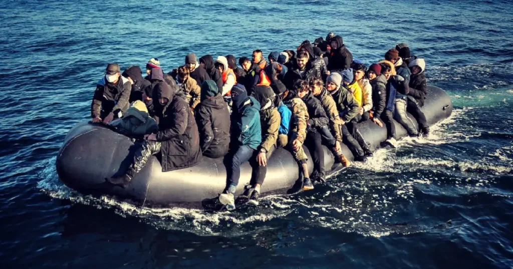 The image shows a group of people on a small inflatable boat in the middle of the ocean, likely indicating a migration or refugee situation.