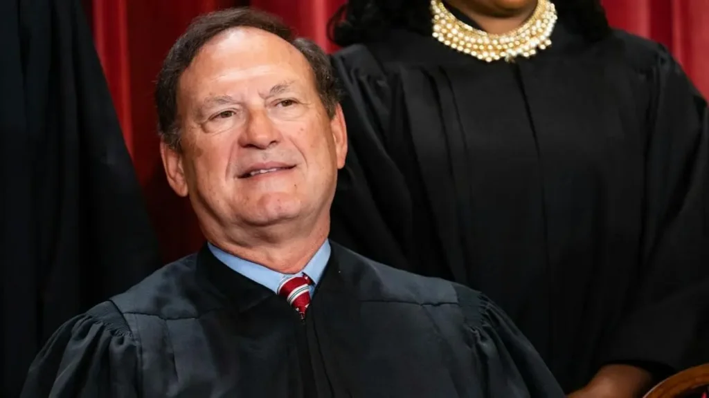 The image shows a person in a judicial robe, seated and smiling, with another individual in a similar robe standing behind them.