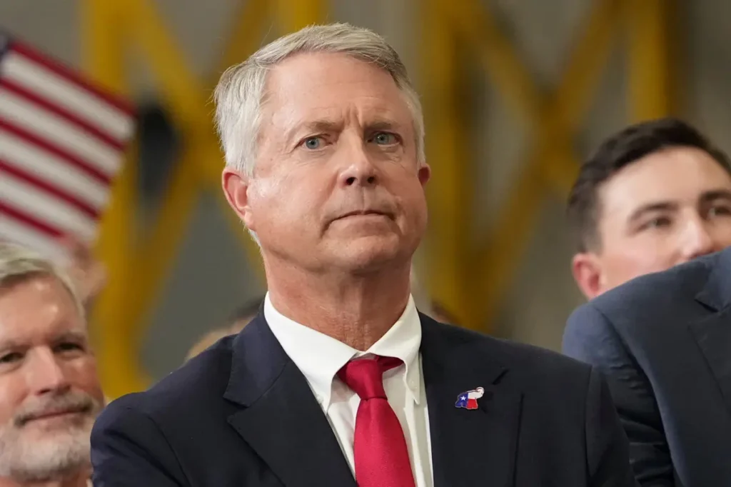 The image shows a man in a suit and tie, wearing a pin with the Texas state flag, seated in a formal setting with other individuals and an American flag in the background.