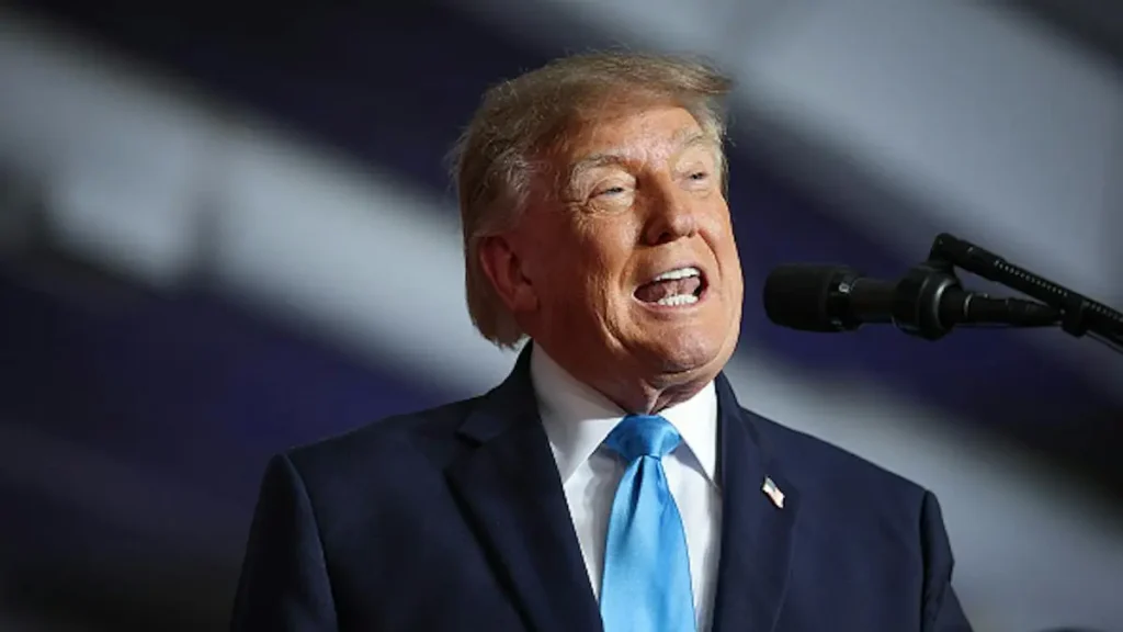 The image shows a person in a suit and tie speaking into a microphone, with a focused and engaged expression.
