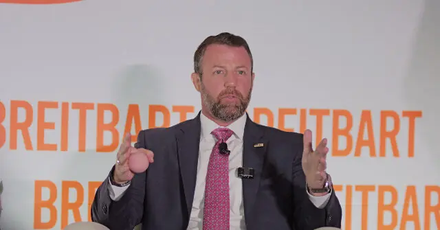 The image shows a man in a suit and tie speaking at a podium with the word "Breitbart" repeated in the background.
