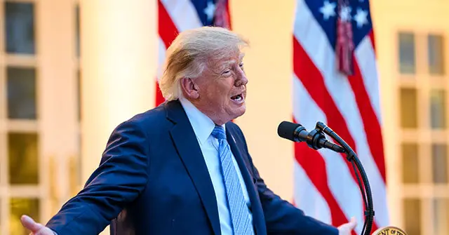 The image shows a person in a suit speaking at a podium with American flags in the background.
