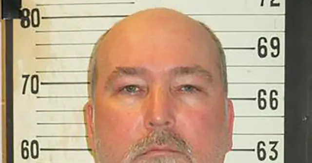 The image is a mugshot of a man with a bald head and a mustache, standing in front of a height chart.