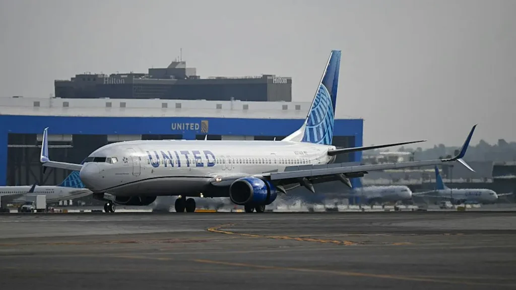 A United Airlines aircraft taxis on the tarmac at an airport, with other planes and airport buildings in the background.