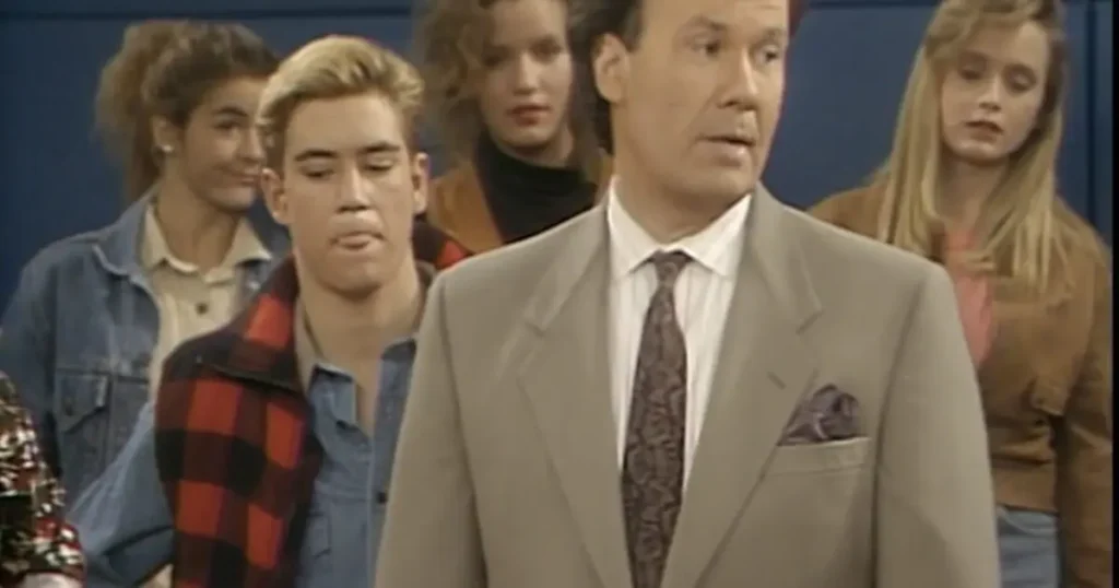 The image shows a man in a suit standing in front of a group of people, possibly in a news or interview setting.