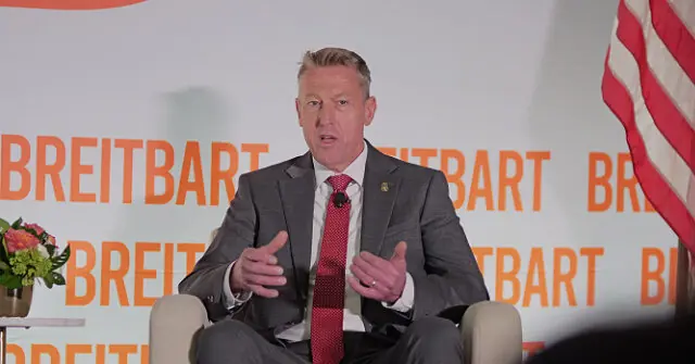 A man in a suit and tie is speaking at a Breitbart event, with an American flag and the Breitbart logo in the background.