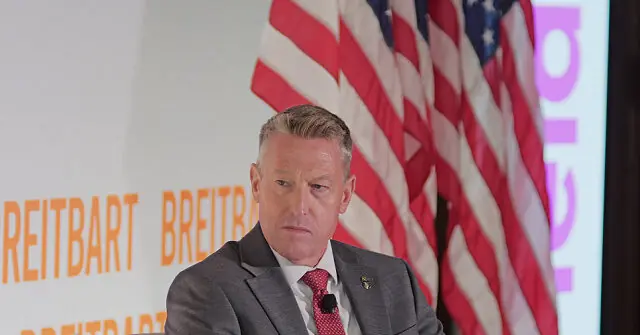 The image shows a man in a suit and tie speaking at a podium with an American flag and the word "Breitbart" in the background.