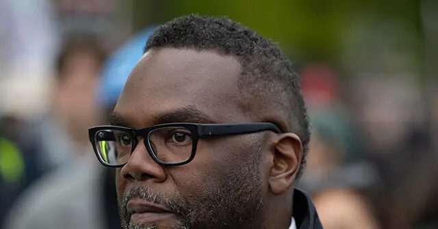 The image shows a close-up of a man wearing glasses, with a blurred background of people and activity.